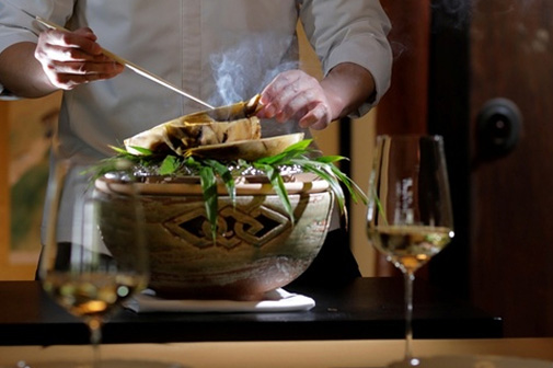 和食の料理人が、土器の器の上で燻される料理を箸で丁寧に盛り付けている様子。手前には白ワインの入ったグラスが置かれている。
